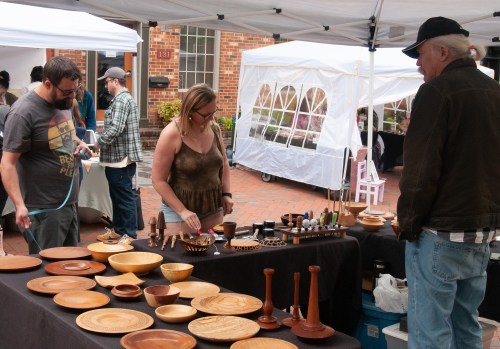 A craft market scene with people browsing wooden items displayed on a table under a tent.