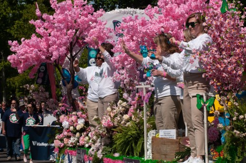 Three people in white shirts wave from a colorful parade float adorned with pink cherry blossoms and flowers.