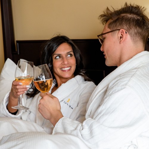 A couple in white robes clinks glasses of wine while sitting in bed, smiling at each other.