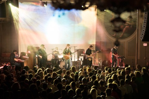 A band performs on stage in front of a large, enthusiastic crowd, with colorful lights and a vibrant backdrop.