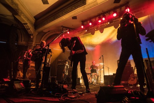 A band performs on stage with dramatic lighting, featuring musicians playing guitar, drums, and other instruments.