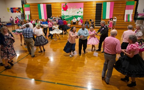 A lively square dance event with couples dancing in colorful outfits in a gym decorated with banners.