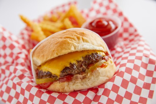 A cheeseburger with pickles and ketchup on a bun, served with fries and a side of ketchup in a red checkered basket.