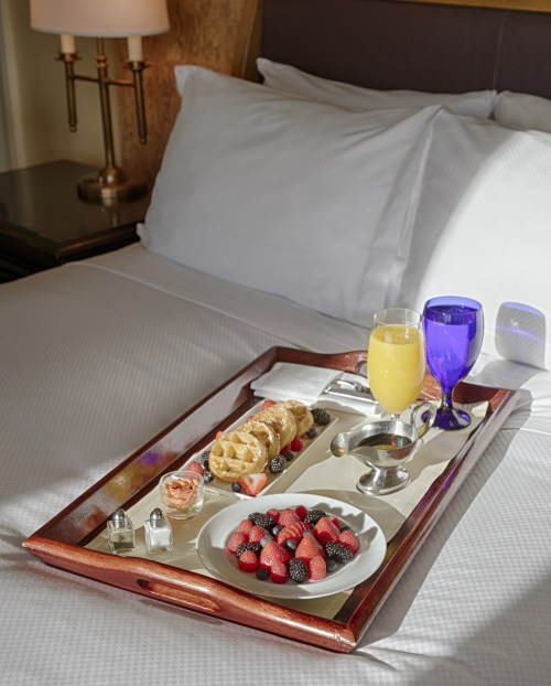 A breakfast tray on a bed featuring waffles, berries, a fruit cup, and drinks in colorful glasses.