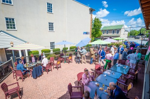 A lively outdoor patio scene with people socializing, enjoying food and drinks under colorful umbrellas on a sunny day.