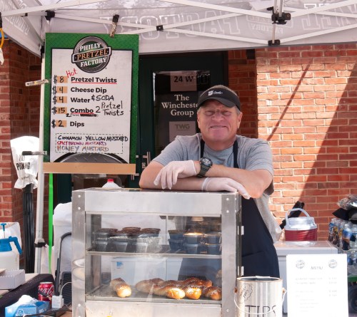 A vendor stands behind a pretzel stand, smiling, with a menu displaying prices for pretzel twists and dips.
