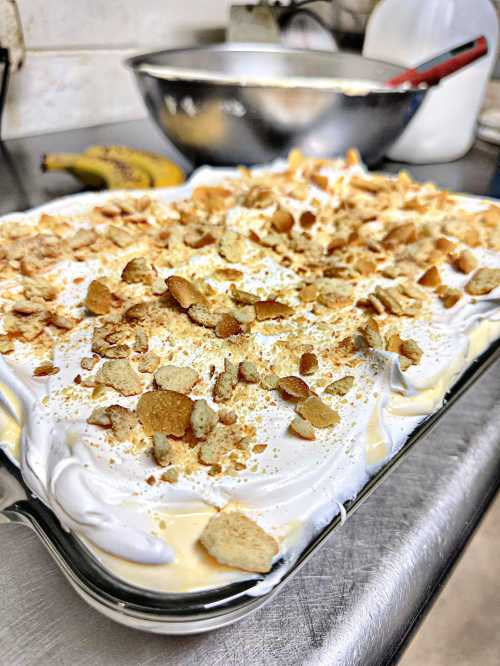 A close-up of a banana pudding dessert topped with whipped cream and crushed cookies, with a mixing bowl in the background.