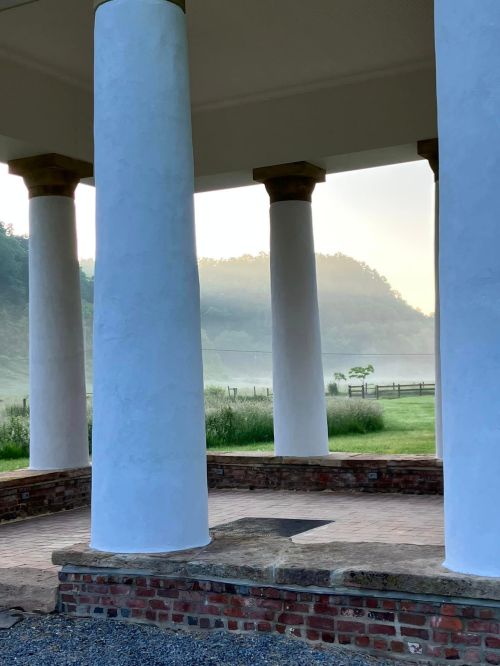 A serene view of white columns supporting a structure, with misty hills and a grassy field in the background.