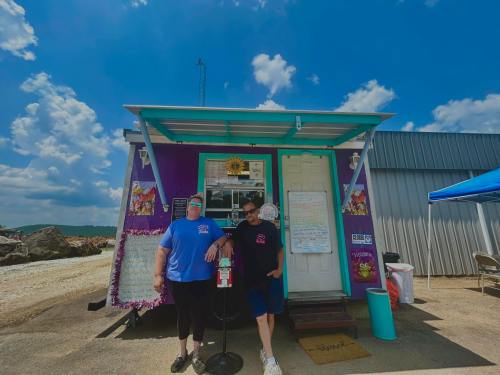 A colorful food truck with two people standing outside, under a bright blue sky with fluffy clouds.