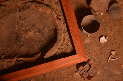An archaeological site featuring a circular stone dish, pottery, and artifacts within a wooden frame on the ground.