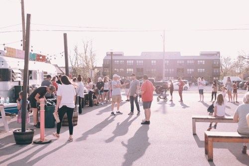 A lively outdoor market scene with people socializing, food trucks, and children playing in the sunlight.