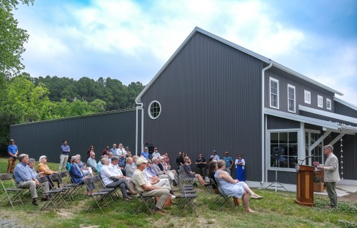 A crowd gathers outdoors for a speech near a modern gray building, with chairs arranged in front of a podium.