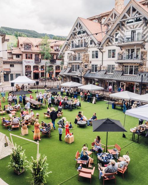 A lively outdoor market scene with people socializing, tents, and greenery in a charming village setting.