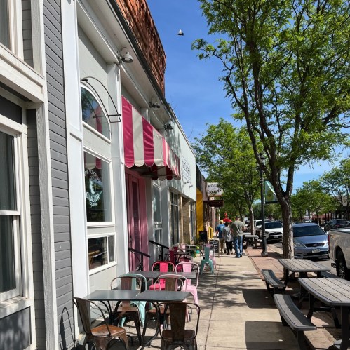 A colorful street scene featuring a café with outdoor seating, trees, and people walking by on a sunny day.
