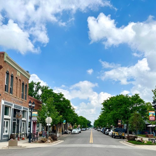 A charming street lined with trees, shops, and parked cars under a blue sky with fluffy clouds.