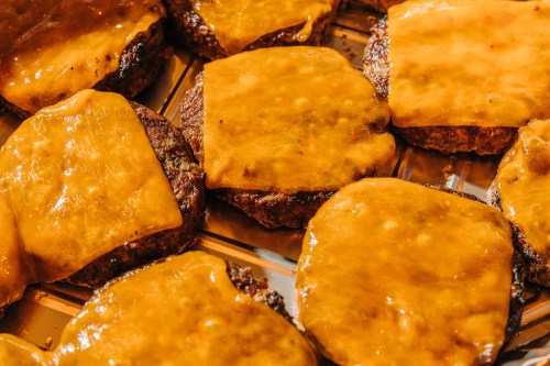 Close-up of grilled beef patties topped with melted cheddar cheese on a baking tray.