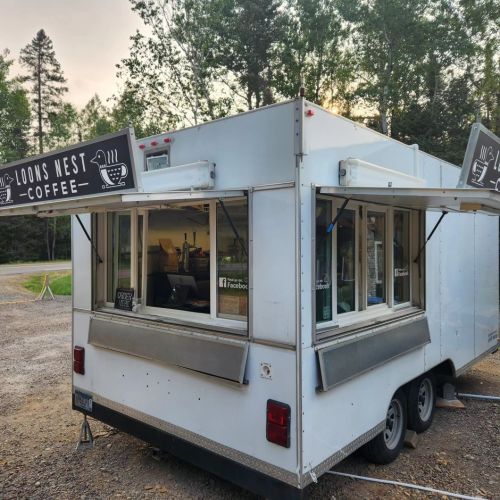 A coffee stand on wheels with a sign reading "Loons West Coffee," surrounded by trees and greenery.