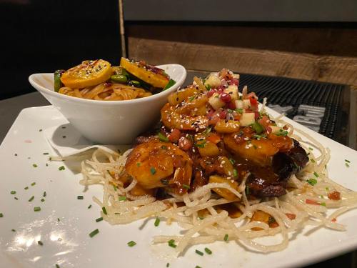 A plate featuring grilled shrimp on a bed of noodles, garnished with vegetables and a side of stir-fried noodles.