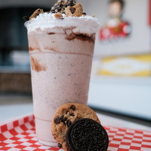 A creamy milkshake topped with whipped cream and cookies, served with a chocolate chip cookie and an Oreo on the side.