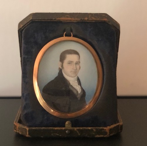 A framed portrait of a man in a black coat, set against a blue background, displayed in a decorative case.