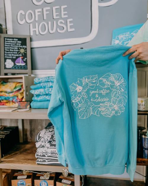A person holds a turquoise sweatshirt with a floral design and the text "Good coffee & kindness" in a coffee shop.