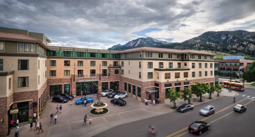 A modern building with a mix of stone and glass, surrounded by trees and mountains, bustling with people and cars.