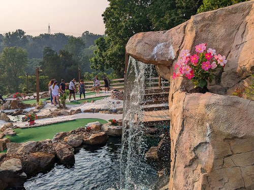 A scenic mini-golf course with a waterfall, flowers, and people enjoying the outdoors in a lush green setting.