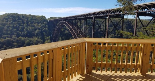 A wooden viewing platform overlooks a lush valley and a large metal bridge spanning the landscape.