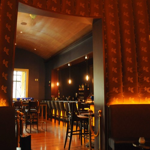 A stylish bar interior with wooden accents, dim lighting, and a row of chairs along a counter.