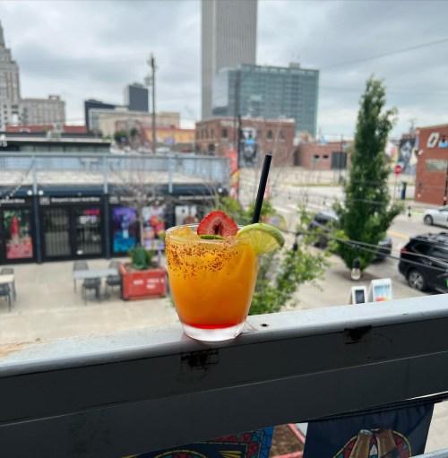 A colorful cocktail with a lime and strawberry garnish sits on a railing, with a cityscape in the background.