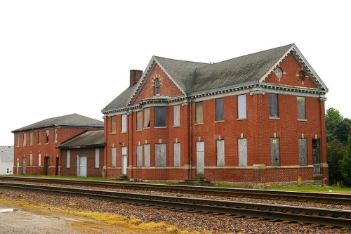 Abandoned red brick buildings with boarded windows near railway tracks, set against a clear sky.