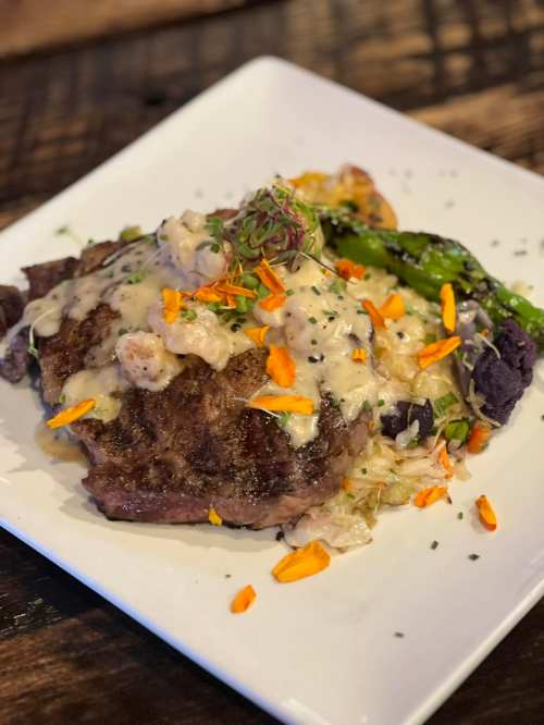 A plated dish featuring a grilled steak topped with creamy sauce, garnished with herbs and colorful edible flowers.