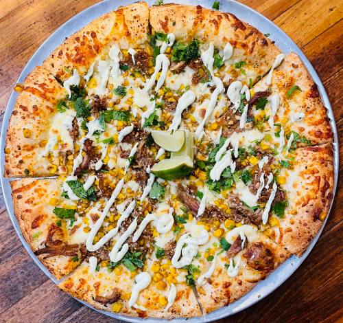 A large pizza topped with meat, corn, herbs, and drizzled with sauce, garnished with lime wedges.