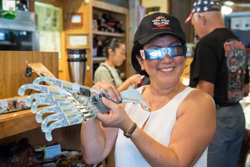 A smiling woman wearing 3D glasses holds novelty sunglasses in a shop, with people browsing in the background.