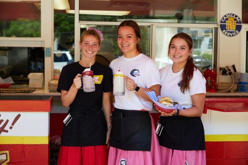 Three young women in matching outfits hold drinks and a food tray at a vibrant outdoor eatery.