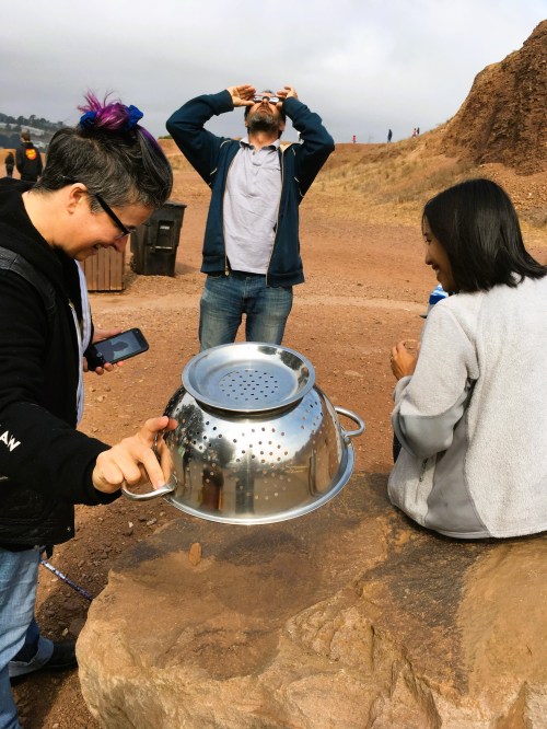 A group of three people outdoors; one pours from a colander, while another drinks and a third looks up.