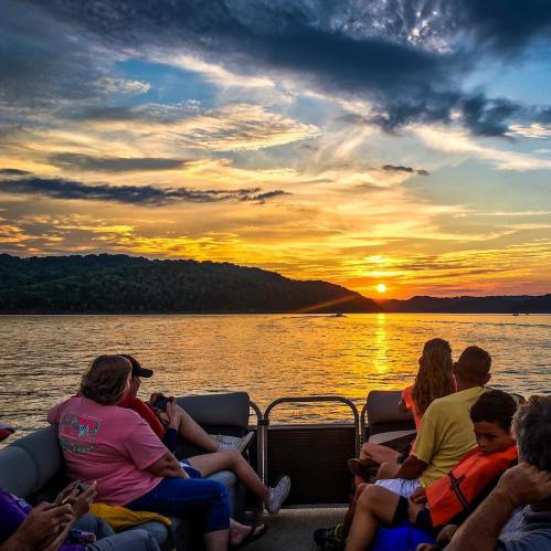 A group of people on a boat watching a vibrant sunset over the water, with hills in the background.