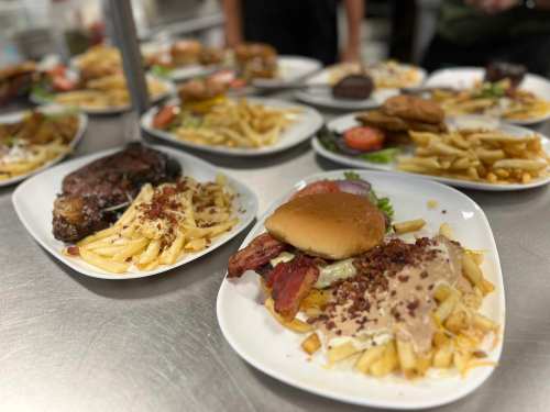 A close-up of a plate with a burger, loaded fries topped with bacon, and various other dishes in the background.