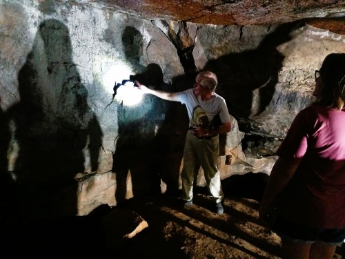 A person points a flashlight at cave wall art while another person observes in a dimly lit cave. Shadows are visible.