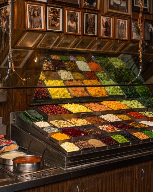 A vibrant display of assorted fruits and vegetables in a market, with framed photos on the wall behind.