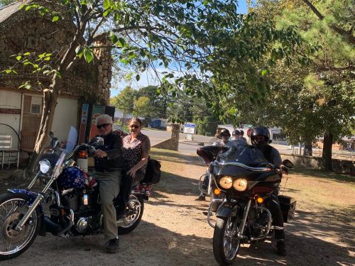 Two motorcycles parked on a dirt path, with riders sitting on them, surrounded by trees and a rustic building.