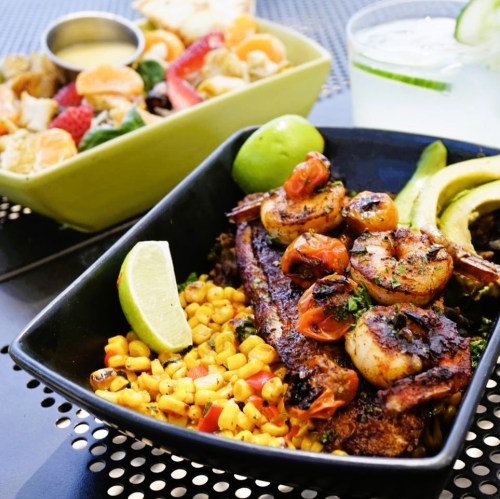 A vibrant dish featuring grilled shrimp and fish on corn, served with lime and a side salad, with a drink in the background.