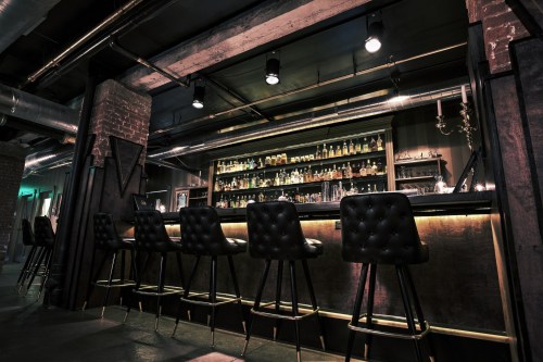 A dimly lit bar with a sleek counter, high stools, and shelves lined with various bottles.