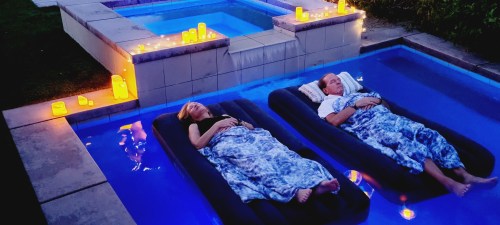 A serene evening scene with two people relaxing on floaties in a pool, surrounded by glowing candles and soft lighting.