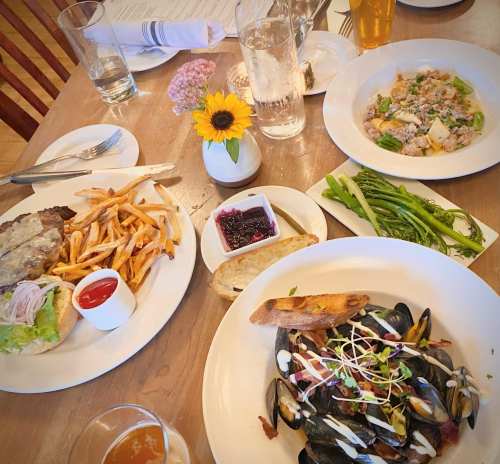 A table set with a burger and fries, mussels, pasta, green beans, and a flower centerpiece. Drinks and condiments are nearby.