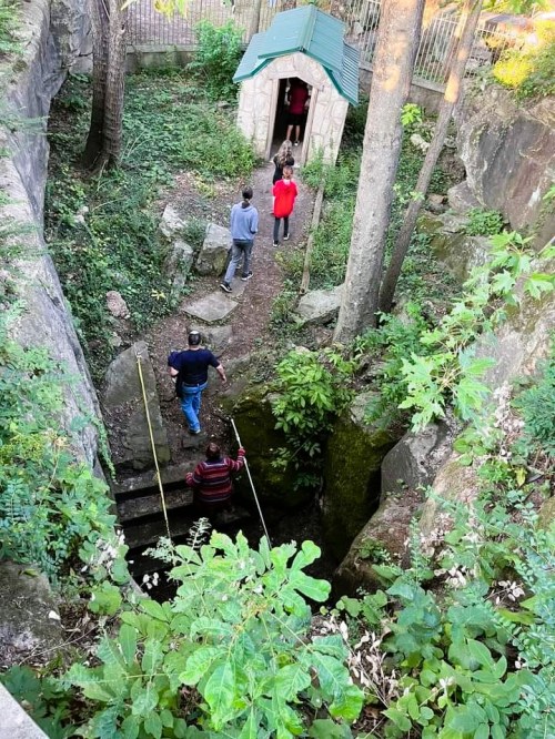 A group of people exploring a rocky area with steps, surrounded by greenery and a small structure in the background.