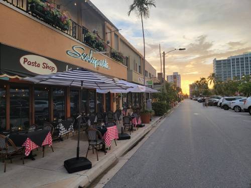 A street view of a pasta shop with outdoor seating, palm trees, and a sunset in the background.