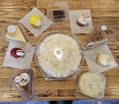 A variety of desserts arranged on a wooden table, including cupcakes, cakes, and slices, all in clear containers.