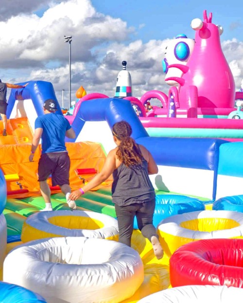 Two people run through a colorful inflatable obstacle course under a cloudy sky, with a large pink character in the background.