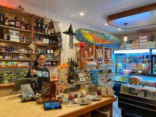 A cozy shop interior with shelves of snacks, drinks, and local products, featuring a smiling cashier at the counter.
